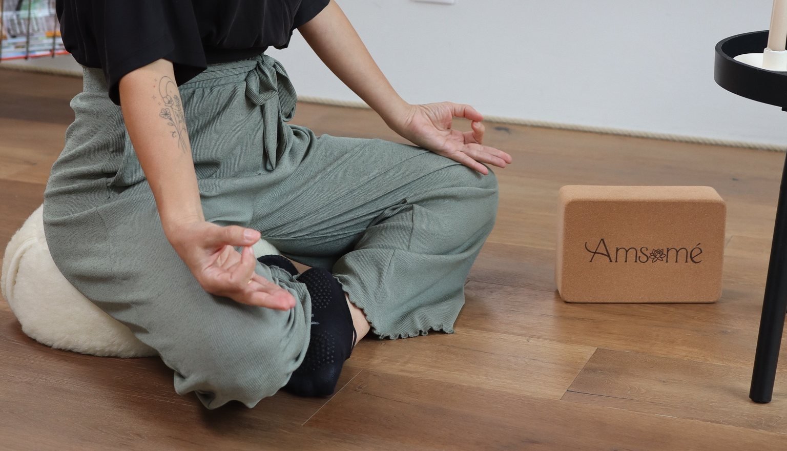 Femme assise en tailleur sur un coussin de méditation en laine, les mains posées sur les genoux, à côté d'une brique de yoga en liège