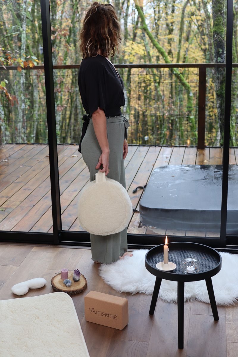 Femme debout tenant un coussin de méditation en laine par sa poignée, devant un espace de méditation avec tapis, bougie et brique
